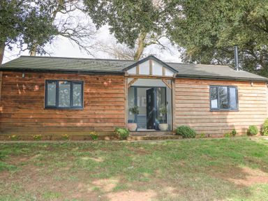 A wooden cottage with two windows and an open front door surrounded by trees at Holm Oaks Cottage in Busbridge near Godalming