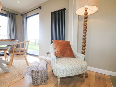A corner of a room with an armchair holding a pillow a basket with blankets a wooden floor lamp and a dining table with chairs near windows at Holm Oaks Cottage in Busbridge near Godalming