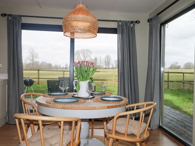 A dining area with a round table set with plates wine glasses and flowers with wooden chairs near large windows at Holm Oaks Cottage Busbridge near Godalming