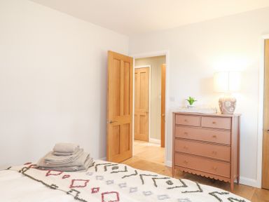 A bedroom with a bed covered with a patterned blanket and towels on top a brown chest of drawers with a lamp and a plant and wooden doors in Holm Oaks Cottage in Busbridge near Godalming
