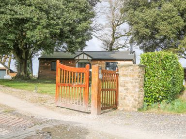 An open wooden gate leading to a driveway with a wooden house and trees in the background at Holm Oaks Cottage in Busbridge near Godalming