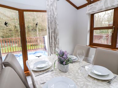 A dining table set with plates glasses cutlery and a flower arrangement in a room with windows showing a wooden deck and outdoor hot tub at 2 Riverside Lodge Abercych near Cenarth