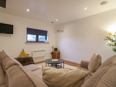 A living room with a beige sofa two cushions a round coffee table a small wooden cabinet a window with a closed blind and a wall-mounted television at Hub House in Three Oaks near Hastings