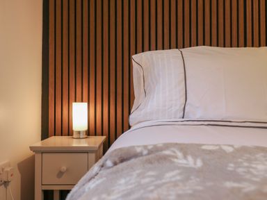A bedside table with a lamp next to a bed with white linens and a wooden slatted headboard at Hub House in Three Oaks near Hastings