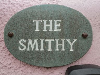 A green oval house name plaque with the text The Smithy on a pink textured wall at The Smithy in Chathill near Seahouses