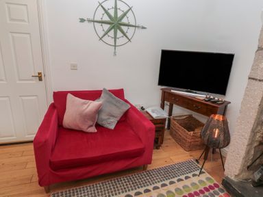 A red armchair with two pillows next to a wooden TV stand with a flat screen TV and a woven basket in a living room at The Smithy in Chathill near Seahouses