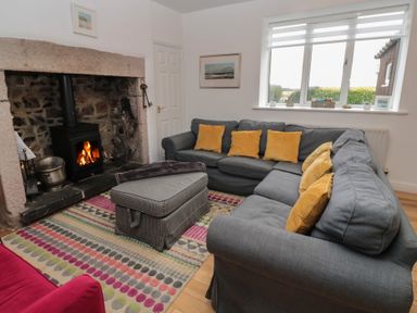 A living room with a stone fireplace and wood stove a gray sectional sofa with yellow cushions and a patterned ottoman on a multi-colored rug at The Smithy in Chathill near Seahouses