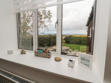 A window sill with decorative items a thermostat and a framed picture overlooking a garden with hedges and a round wooden table at The Smithy in Chathill near Seahouses