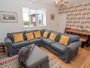 A living room with a gray sectional sofa and yellow cushions a wooden dining table with chairs and patterned wallpaper at The Smithy in Chathill near Seahouses