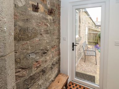 A hallway with an exposed stone wall a wooden bench and a glass door leading outside to a gravel area with a metal bench at The Smithy in Chathill near Seahouses