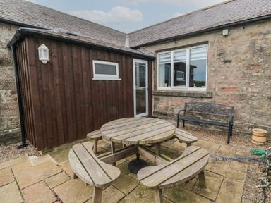 An outdoor patio with a round wooden picnic table and benches near a stone wall and a wooden shed at The Smithy in Chathill near Seahouses
