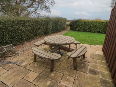 A wooden round picnic table with attached benches on a stone patio beside a lawn and hedge