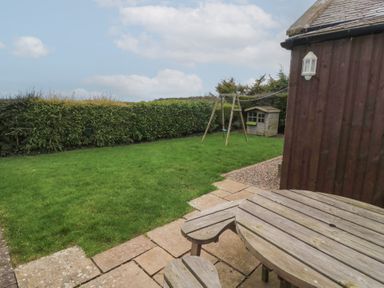 A garden with a grass lawn a wooden swing a small wooden playhouse and a round wooden picnic table at The Smithy in Chathill near Seahouses