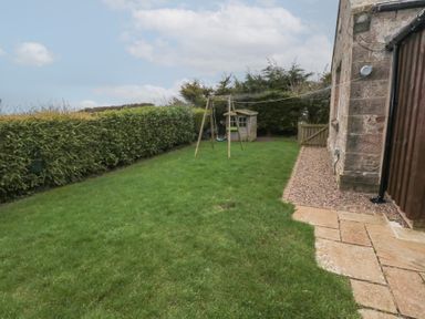 A backyard with grass lawn swing set playhouse and stone pathway next to a stone building at The Smithy in Chathill near Seahouses