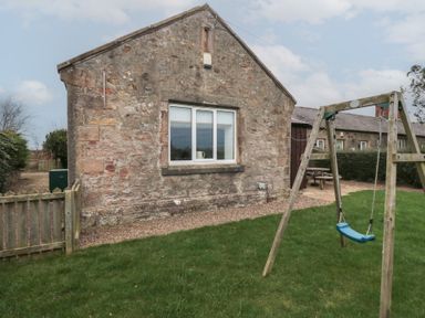 A stone building with a window and a swing set on a grassy lawn at The Smithy in Chathill near Seahouses