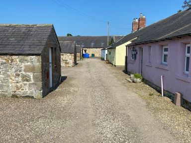 A gravel pathway between stone and pastel-colored buildings with chimneys under a clear sky at The Smithy in Chathill near Seahouses