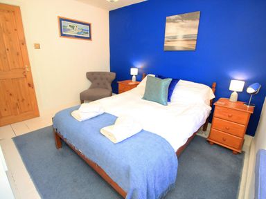 A bedroom with a double bed blue and white bedding two wooden bedside tables with lamps and a gray chair by a wooden door at Riverbank Cottage in Beddgelert