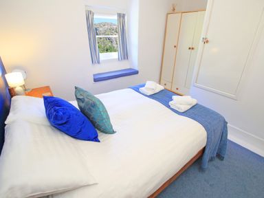 A bedroom with a bed blue pillows folded towels wooden wardrobe and window seat at Riverbank Cottage in Beddgelert