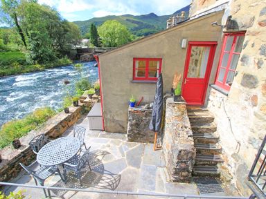 An outdoor patio area with a round metal table and four chairs next to a flowing river and a stone building with red window frames and door at Riverbank Cottage in Beddgelert