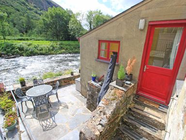 An outdoor patio with metal table and chairs next to a river and stone steps leading to a red door at Riverbank Cottage in Beddgelert