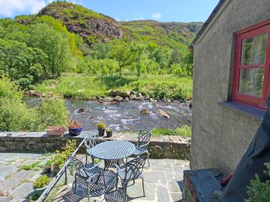 An outdoor patio with a metal table and four chairs next to a river and green hillside at Riverbank Cottage in Beddgelert