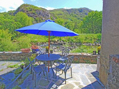 An outdoor patio with a blue umbrella, metal table and chairs by a stone wall next to a river and hills at Riverbank Cottage in Beddgelert
