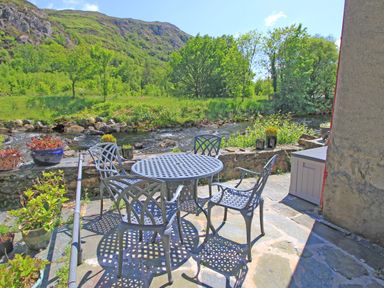 An outdoor patio with metal table and chairs by a river and trees at Riverbank Cottage in Beddgelert