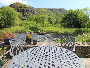 An outdoor patio with metal table and chairs overlooking a river and green hillside at Riverbank Cottage in Beddgelert