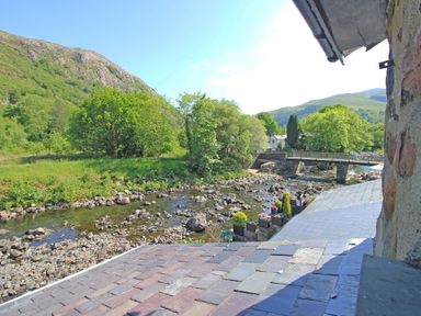 A river with rocks along the bank and a bridge in a green valley with hills and trees at Riverbank Cottage in Beddgelert