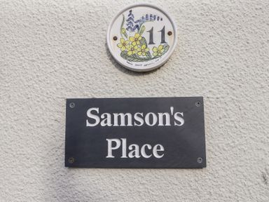 A wall with a house number 11 plaque decorated with flowers and a black sign reading Samsons Place at Morfa Bychan