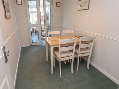 A dining area with a wooden table and four white chairs with cushions in a narrow room at Samson's Place Morfa Bychan