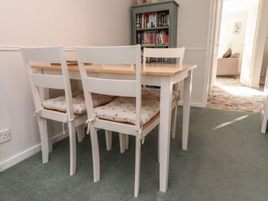 A dining area with a wooden table and four white chairs with cushions a bookshelf filled with books and a view into a living room at Samson's Place in Morfa Bychan