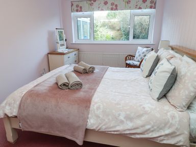 A bedroom with a double bed covered in floral bedding and pillows with text a small dresser with mirror near the window and a chair next to the bed at Samsons Place in Morfa Bychan
