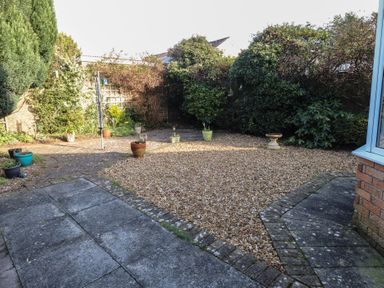 A backyard with gravel area and stone patio surrounded by bushes and potted plants at Samson's Place in Morfa Bychan