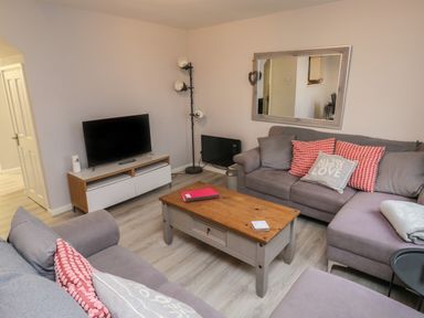 A living room with a gray sectional sofa with patterned cushions a wooden coffee table a television on a stand a floor lamp and a large mirror on the wall at Lewis Lodge in St Ann's Chapel