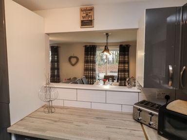 A kitchen countertop with a toaster and microwave and a window opening to a dining area with a table set and plaid curtains at Lewis Lodge in St Ann's Chapel