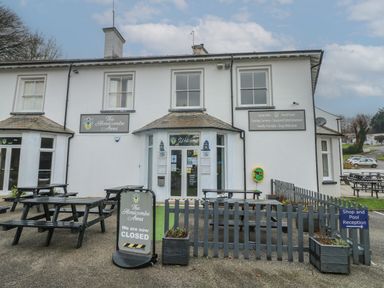 Exterior view of a building with signs for The Honicombe Arms and outdoor picnic tables in front at Lewis Lodge in St Ann's Chapel
