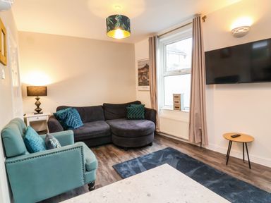 A living room with a dark grey corner sofa green armchair wall mounted tv and window with curtains at Hollycombe in Ilfracombe