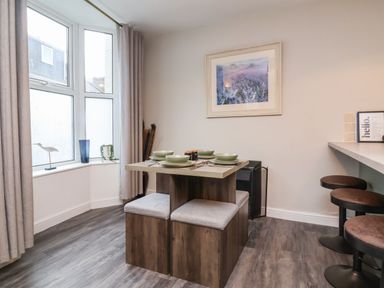 A dining area with a wooden table set for four and cushioned stools near a window with curtains at Hollycombe in Ilfracombe