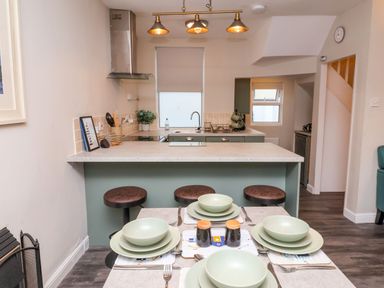 A kitchen with a countertop and three bar stools and a dining table set with plates bowls and cutlery at Hollycombe in Ilfracombe