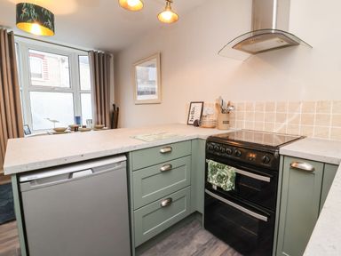 A kitchen with green cabinets, a black stove, a silver dishwasher, a tiled backsplash, and a dining area with bowls near a window at Hollycombe in Ilfracombe