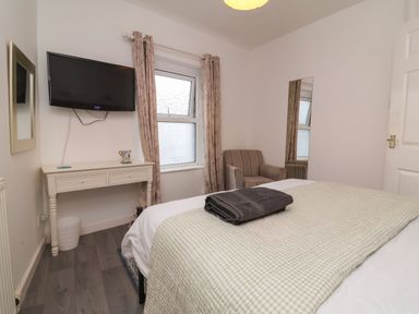 A bedroom with a bed covered with a blanket and folded towel a wall-mounted television a window with curtains a small desk a striped armchair and a wall mirror at Hollycombe in Ilfracombe