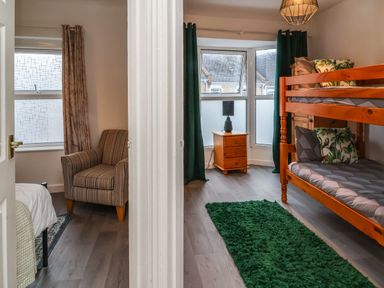 A room with a wooden bunk bed pillows green curtains and a wooden nightstand with a black lamp and an adjacent room with a striped armchair and a window at Hollycombe in Ilfracombe
