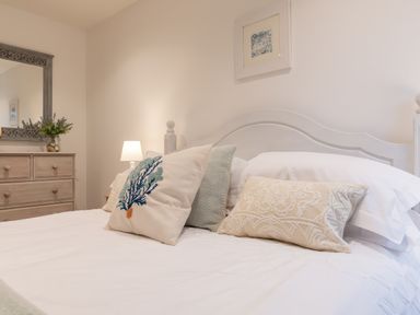 A bedroom with a white bed with decorative pillows a wooden dresser with a mirror and a table lamp at Oystercatcher in Mevagissey