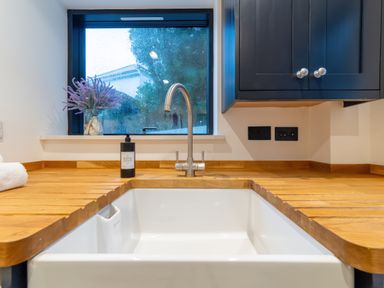 A kitchen sink with a faucet and hand soap on a wooden countertop under a window with a flower vase at Oystercatcher in Mevagissey