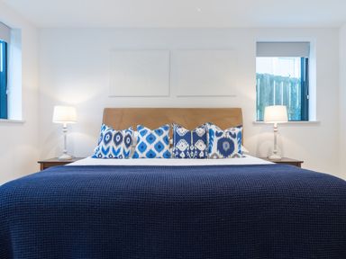 A bedroom with a bed with blue patterned pillows and a navy blanket between two windows with lamps on bedside tables at Oystercatcher in Mevagissey