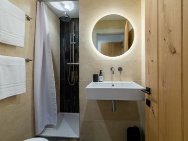 A bathroom with a shower area a white sink and a round illuminated mirror at Oystercatcher Mevagissey