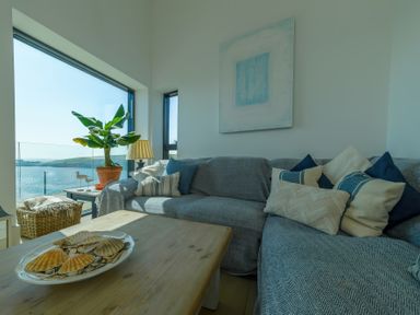 A living room with a sofa pillows coffee table and a plant by large windows overlooking the sea at Oystercatcher in Mevagissey