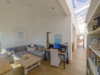 A living area with a gray sectional sofa and coffee table next to a desk with a computer and chair and a bookshelf in a hallway at Oystercatcher in Mevagissey