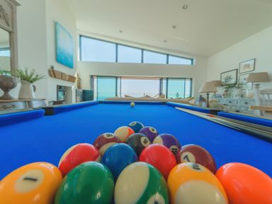 A pool table with balls racked and cues resting on the side in a room with large windows and a seating area at Oystercatcher in Mevagissey